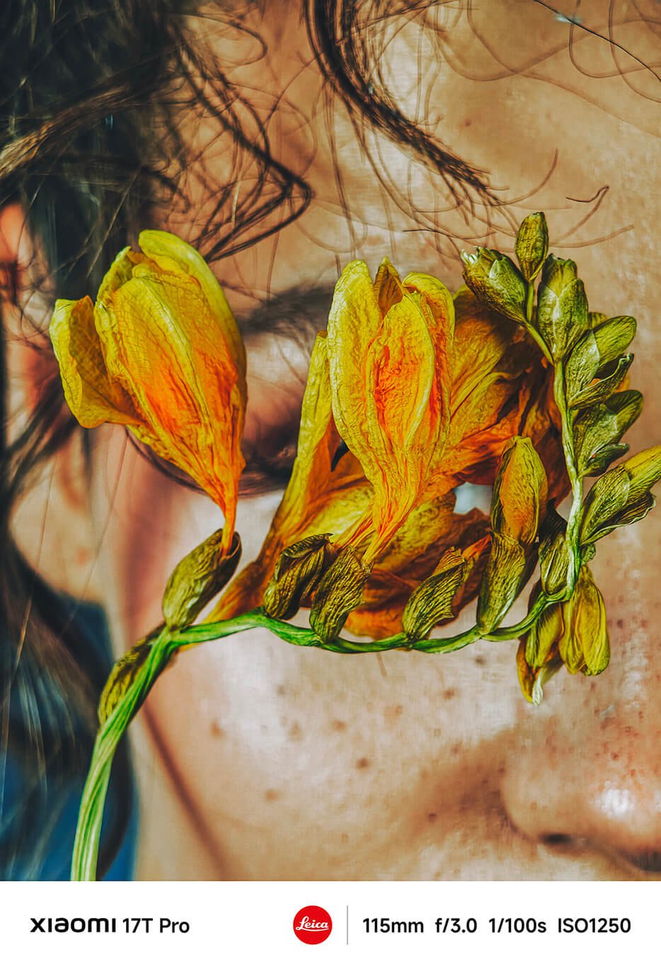 Un buchet de flori galbene acoperă fața unei persoane, iar fundalul este neclar, evidențiind nuanțele delicate ale pielii și freckle-urile. Forma și culoarea florilor contrastează cu fața, creând un efect vizual unic.