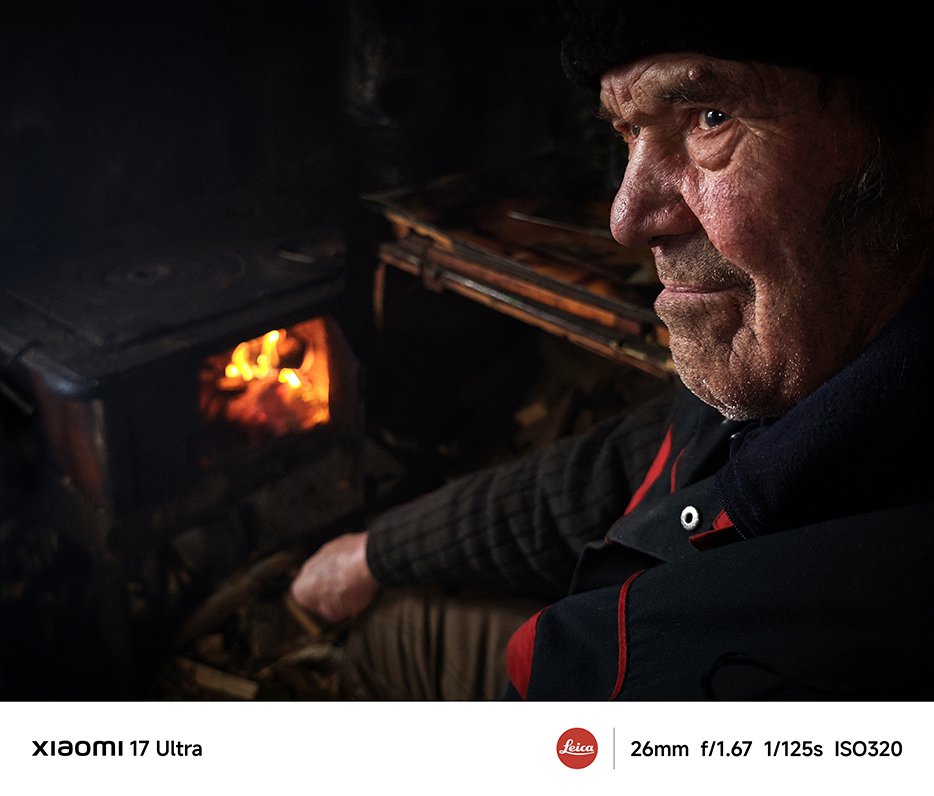 Un bătrân stă lângă sobă, cu o expresie adânc gânditoare pe față, flăcările din sobă strălucesc, iar fundalul este mai întunecat, creând o atmosferă caldă și liniștită.