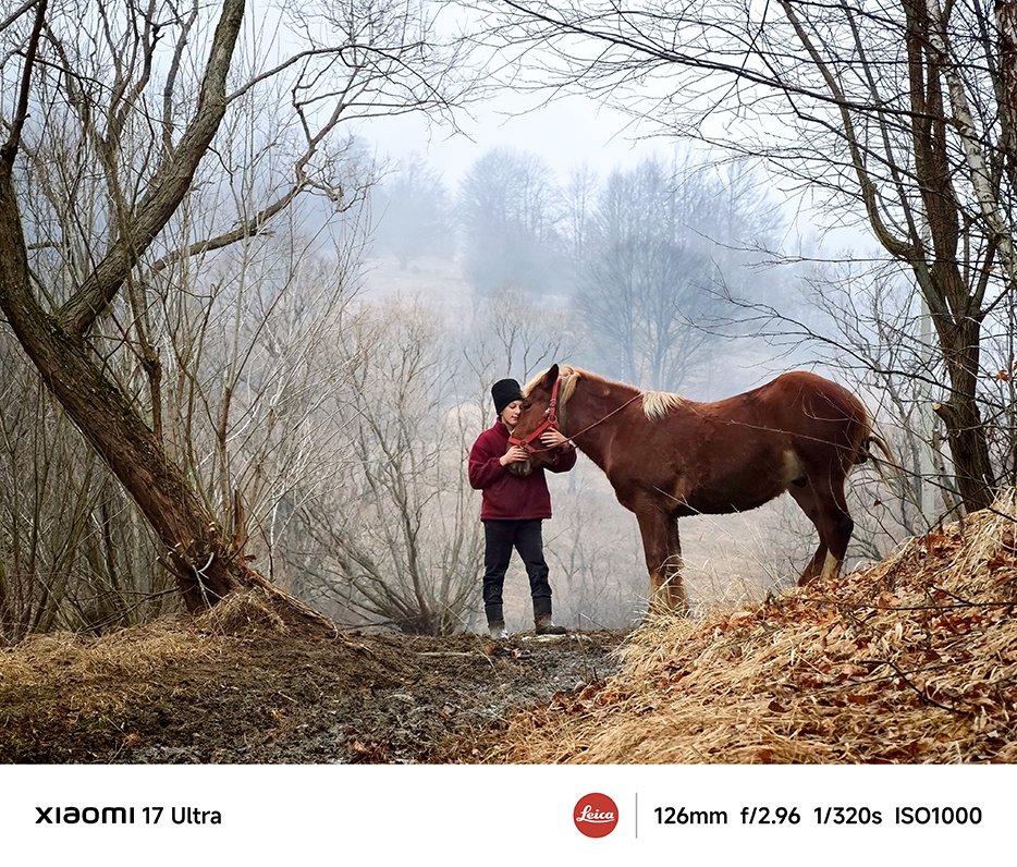 Un om stă în pădure, mângâind un cal brun. În jur sunt copaci golași și iarbă uscată, iar în aer plutește o ceață ușoară.