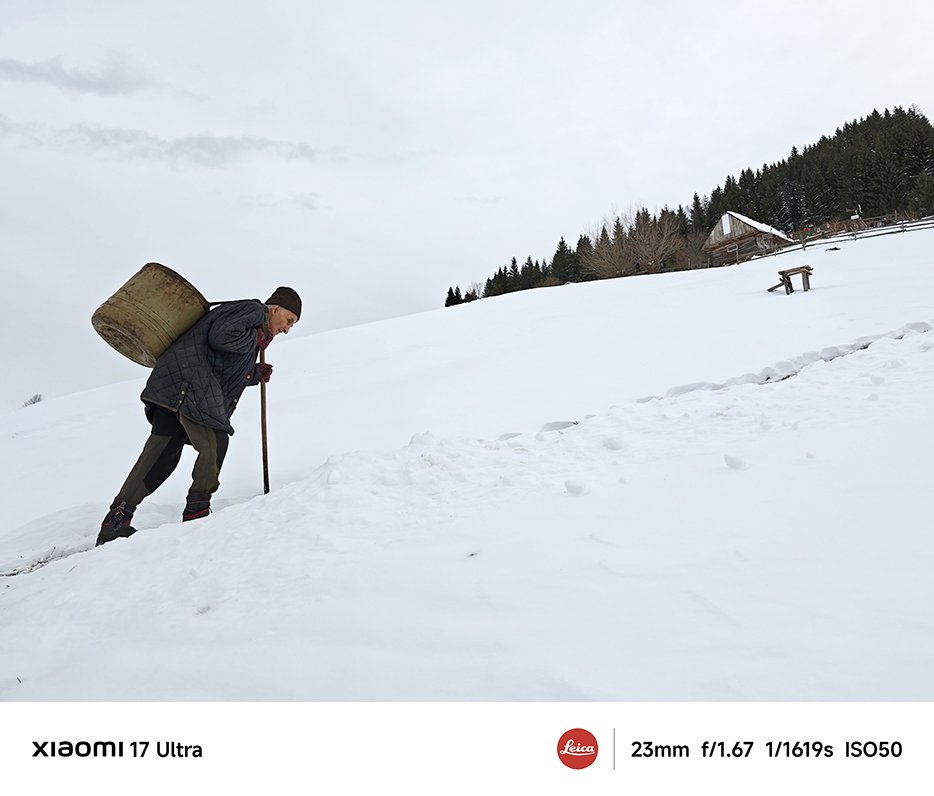 Un trecător cu un butoi rotund în spate merge pe zăpadă, iar lângă el se află pământ și copaci acoperiți cu zăpadă, departe se poate vedea o mică casă. Întregul mediu pare rece și liniștit.