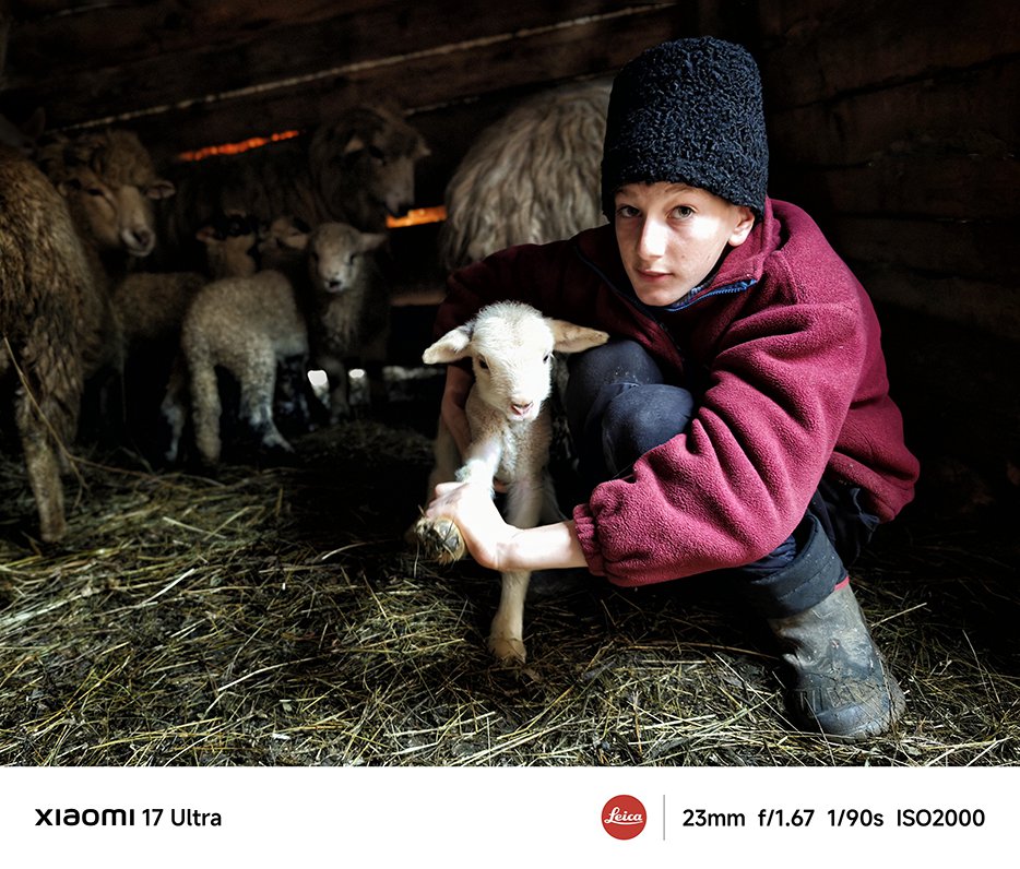 O persoană îmbrăcată într-o haină roșie ține în brațe un miel în staul, înconjurată de alte oi, iar mediul pare cald și liniștit.